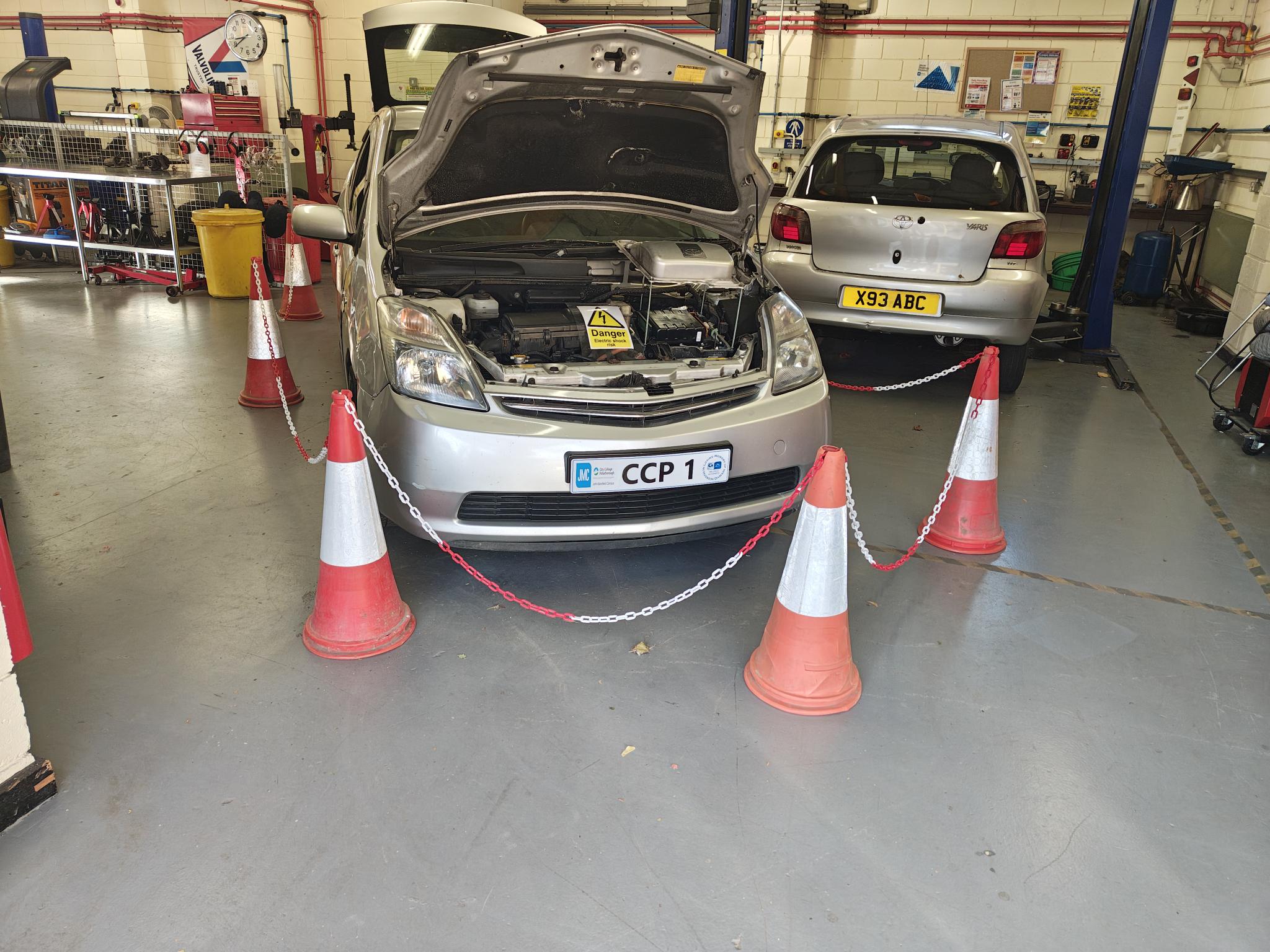 College workshop bay with a Toyota Prius hybrid, bonnet open, with safety cones and chain in place; another project car in the background.
