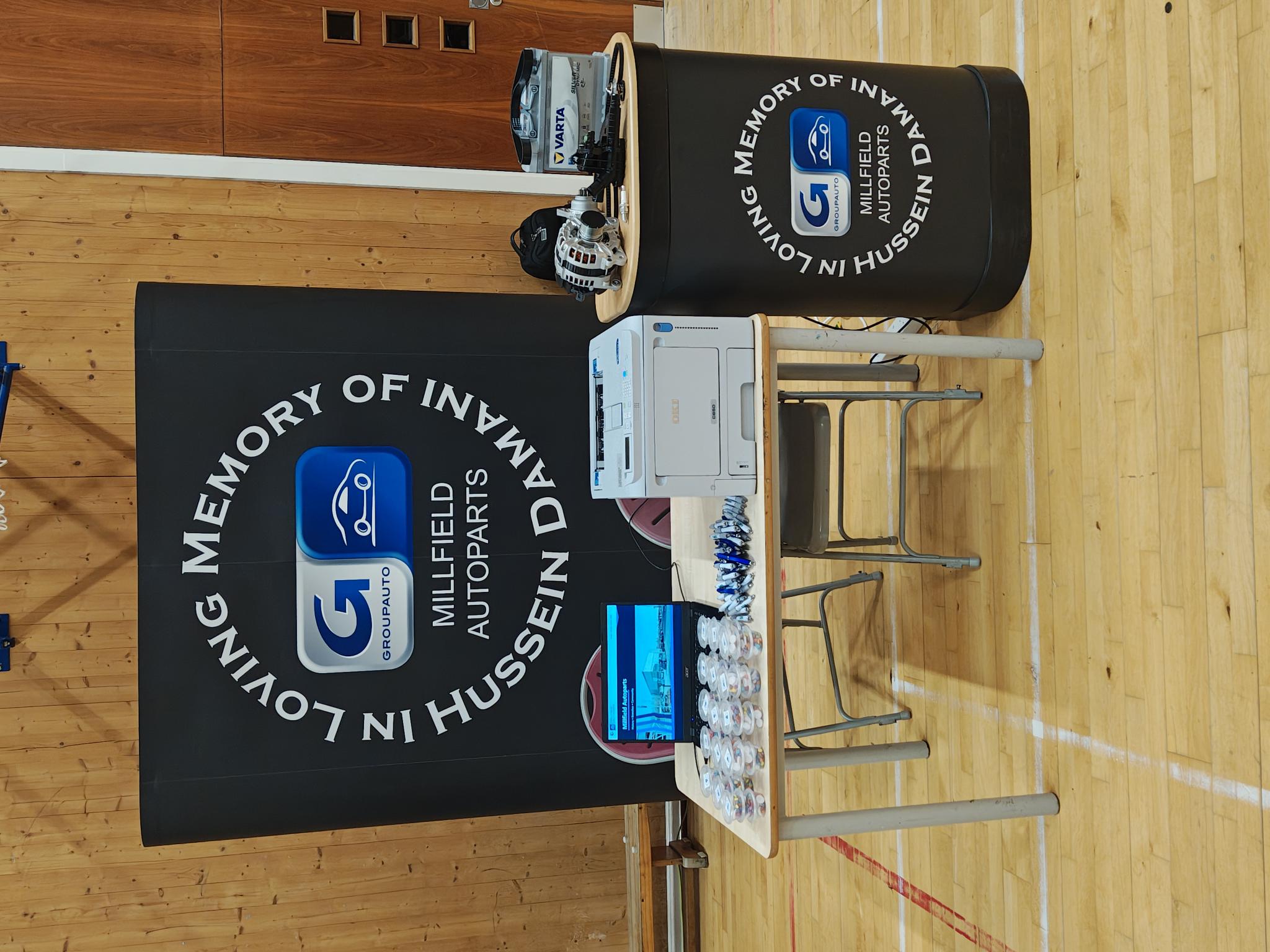 Millfield Auto Parts stand at St John Fisher Catholic High School careers fair with memorial banner, laptop, printer, giveaways, alternator and Varta battery on display.